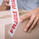 Ręce zaklejają karton taśmą z napisem "FRAGILE". Uwaga szkło kurier musi być ostrożny.