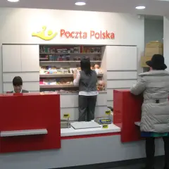 W placówce Poczty Polskiej, pracownica obsługuje klienta. W tle półki z produktami, a obok stos paczek. To miejsce, gdzie realizowane są usługi global expres.