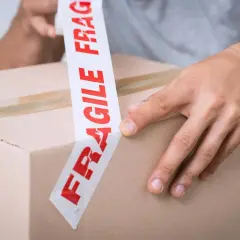 Ręce zaklejają karton taśmą z napisem "FRAGILE". Uwaga szkło kurier musi być ostrożny.