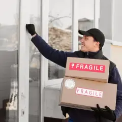 Kurier z paczkami z napisem "FRAGILE" puka do drzwi, realizując wysyłkę szyby samochodowej kurierem.