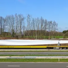Żółta ciężarówka transportuje ogromne łopaty wiatraka, które są **ładunkami nienormatywnymi**. Długi, biały element spoczywa na specjalnej platformie.