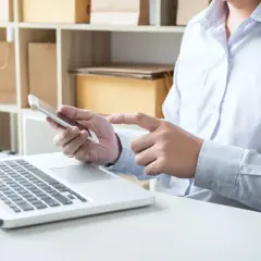 Osoba sprawdza na telefonie, czy otrzymano dane elektroniczne przesyłki, obok leży laptop. W tle regał z paczkami.