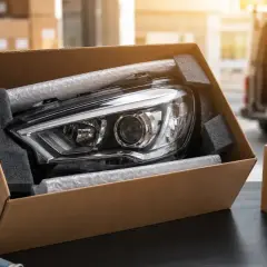 Nowy reflektor samochodowy zabezpieczony do transportu części samochodowych. Obok kartony i taśma klejąca.