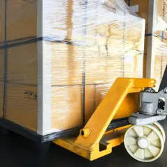 Wózek paletowy przygotowuje transport materaca. Duże, owinięte folią pudła czekają na załadunek.