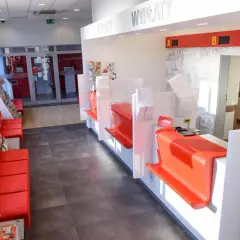 Wnętrze poczty z czerwonymi kanapami i regałami z prasą. Okienka do obsługi klienta z tabliczkami "WPŁATY" i "WYPŁATY". Zcpp zwolnienie przesyłki do doręczenia.