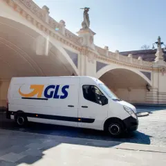 Biały van GLS z logo firmy jedzie pod łukowym mostem, dostarczając paczkę.