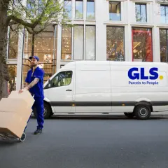 Kurier GLS z paczkami na wózku przed białym vanem. Dowiedz się, jak nadać paczkę z GLS.