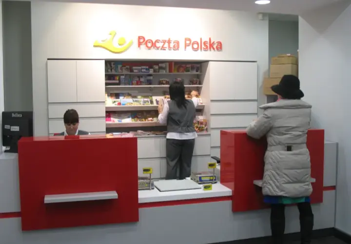 W placówce Poczty Polskiej, pracownica obsługuje klienta. W tle półki z produktami, a obok stos paczek. To miejsce, gdzie realizowane są usługi global expres.