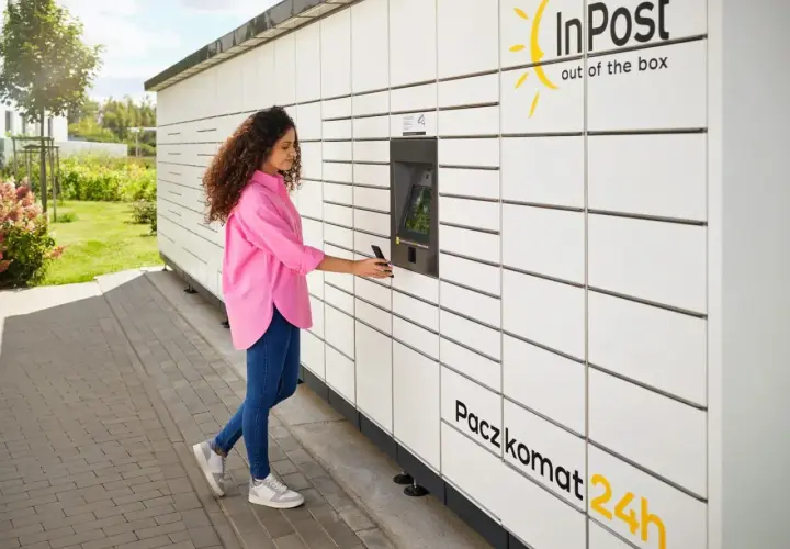 Kobieta z telefonem przy Paczkomacie InPost. Czy z paczkomatu można wysłać paczkę do domu? Tak, to wygodne rozwiązanie.