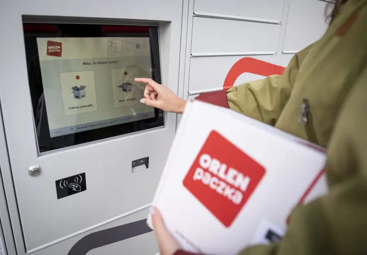 Kobieta z paczką ORLEN PACZKA dotyka ekranu automatu, wybierając opcję nadania przesyłki.
