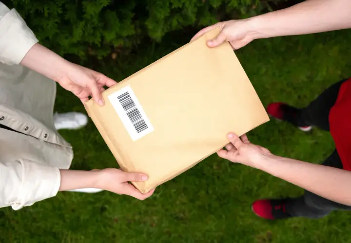 Dwie osoby przekazują sobie paczkę. Jedna z nich ma na rękawie białą koszulę, druga czerwoną bluzkę. Możliwe, że to anulowanie paczki InPost.