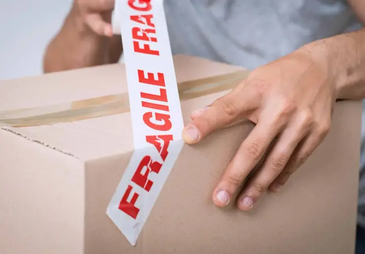 Ręce zaklejają karton taśmą z napisem "FRAGILE". Uwaga szkło kurier musi być ostrożny.