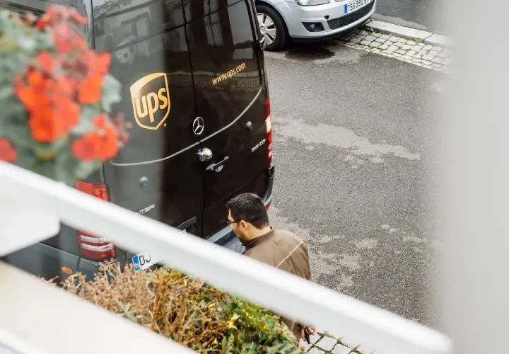 Kurier UPS z paczką zastanawia się, ups gdzie nadać. Obok stoi srebrny samochód.