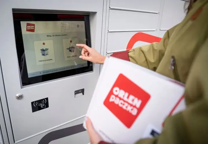 Dłoń dotyka ekranu automatu paczkowego, wybierając opcję "Nadaj przesyłkę". W ręku trzymana jest biała paczka z logo ORLEN paczka.