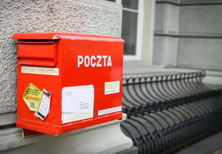 Czerwona skrzynka pocztowa z napisem "POCZTA" i naklejkami. Gotowa na przesyłkę do Wielkiej Brytanii, z adresem i kodem pocztowym.