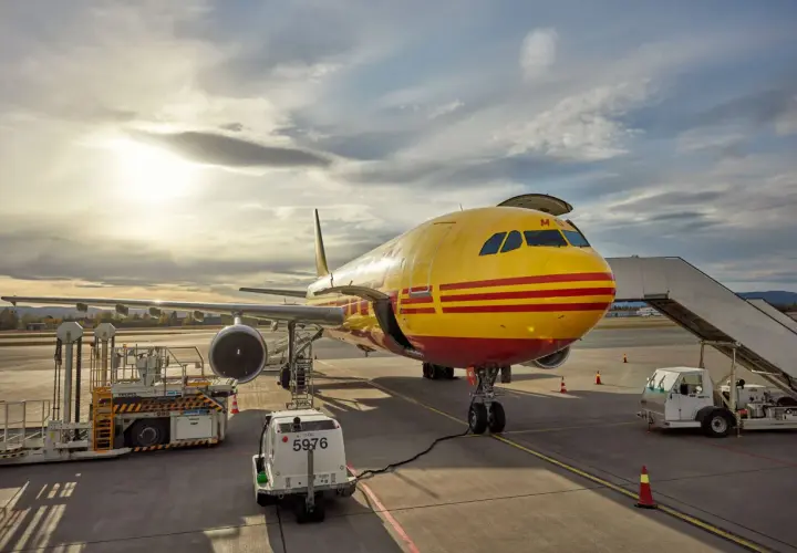 Żółty samolot DHL z czerwonymi pasami na płycie lotniska, gotowy do lotu do USA.