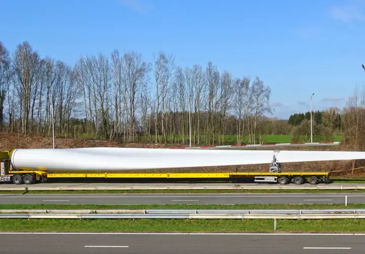 Żółta ciężarówka transportuje ogromne łopaty wiatraka, które są **ładunkami nienormatywnymi**. Długi, biały element spoczywa na specjalnej platformie.