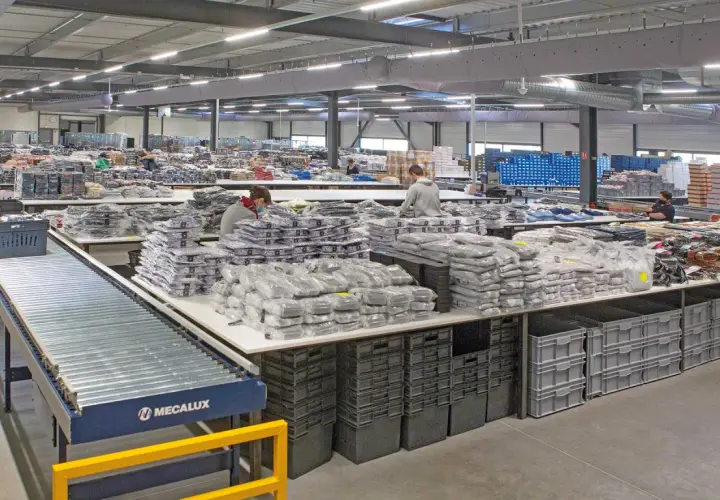 Pracownicy sortują odzież w magazynie. Widać taśmociąg, stoły z zapakowanymi produktami i pojemniki. To pokazuje różne funkcje magazynu.