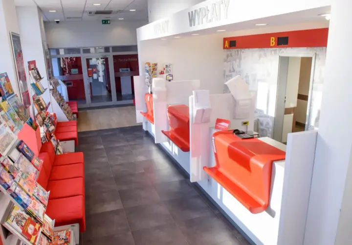 Wnętrze poczty z czerwonymi kanapami i regałami z prasą. Okienka do obsługi klienta z tabliczkami "WPŁATY" i "WYPŁATY". Zcpp zwolnienie przesyłki do doręczenia.
