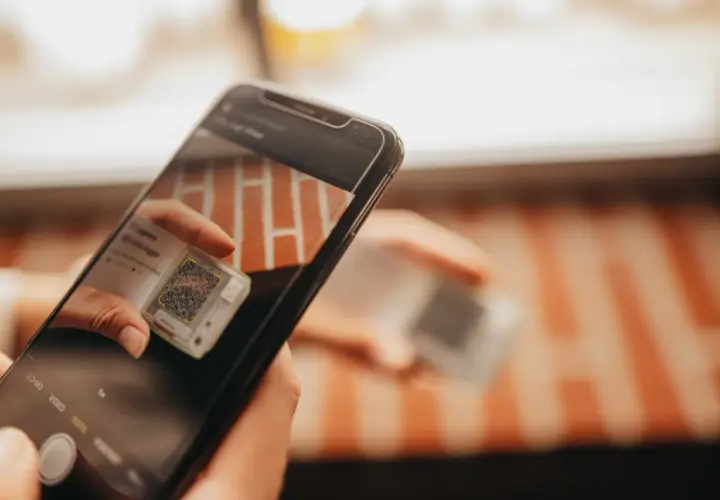 Ręce trzymają smartfon, który skanuje kod QR na małym urządzeniu. To może być część procesu śledzenia przesyłki przez kuriera Hellmann.