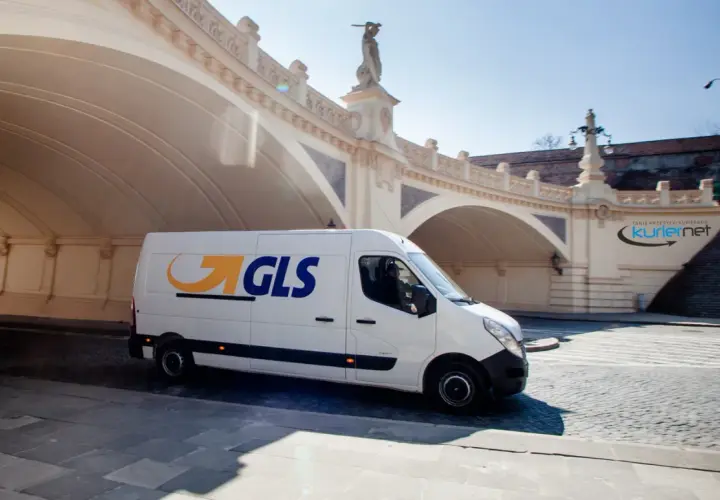 Biały van GLS z logo firmy jedzie pod łukowym mostem, dostarczając paczkę.