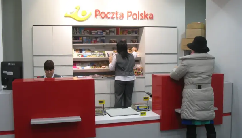 W placówce Poczty Polskiej, pracownica obsługuje klienta. W tle półki z produktami, a obok stos paczek. To miejsce, gdzie realizowane są usługi global expres.