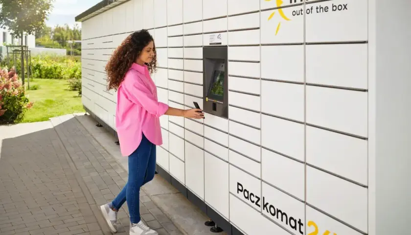 Kobieta z telefonem przy Paczkomacie InPost. Czy z paczkomatu można wysłać paczkę do domu? Tak, to wygodne rozwiązanie.