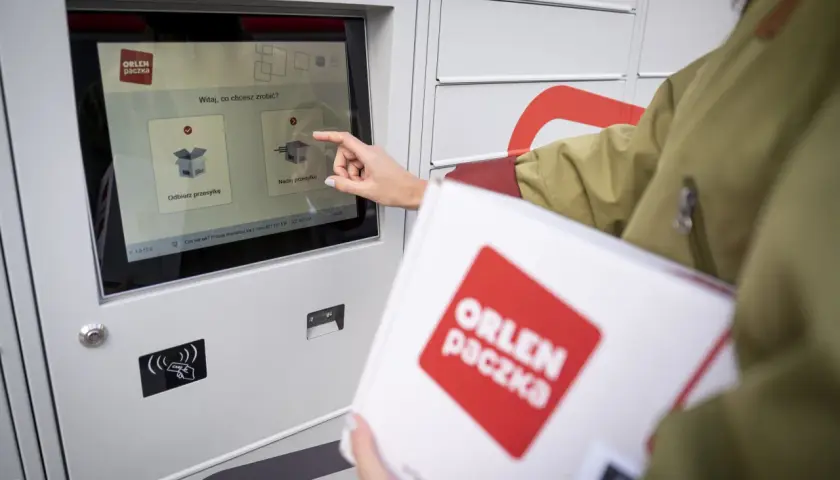 Kobieta z paczką ORLEN PACZKA dotyka ekranu automatu, wybierając opcję nadania przesyłki.