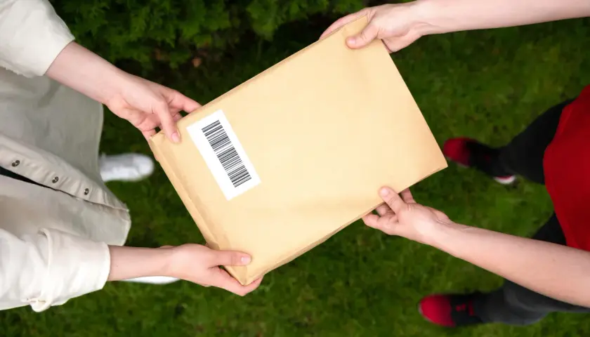 Dwie osoby przekazują sobie paczkę. Jedna z nich ma na rękawie białą koszulę, druga czerwoną bluzkę. Możliwe, że to anulowanie paczki InPost.