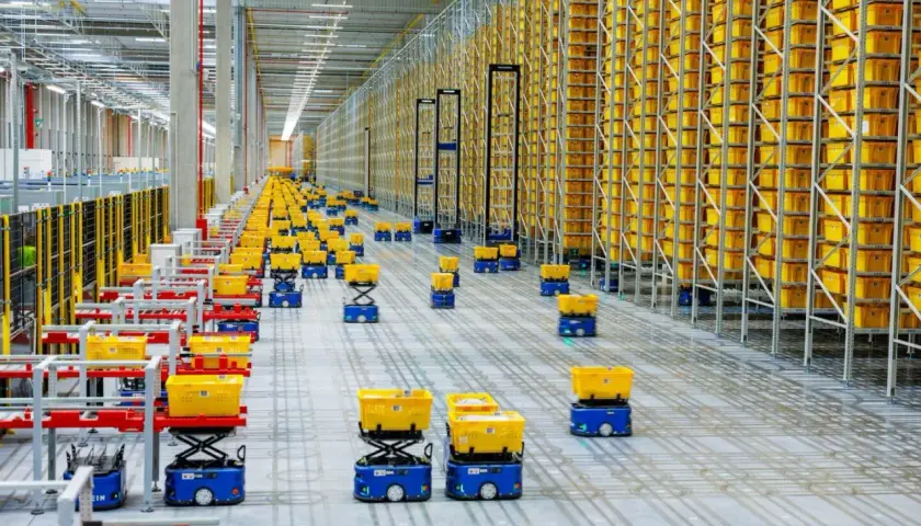 Nowoczesny hub logistyczny z autonomicznymi robotami przewożącymi żółte kosze pośród wysokich regałów wypełnionych towarami.
