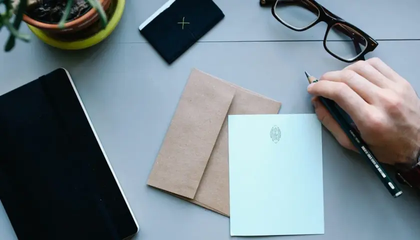Ręka z ołówkiem przygotowuje się do napisania listu. Obok leżą koperta i papeteria, idealne, by dowiedzieć się, jak zaadresować list do Anglii.