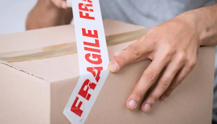 Ręce zaklejają karton taśmą z napisem "FRAGILE". Uwaga szkło kurier musi być ostrożny.