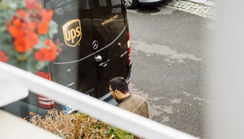Kurier UPS z paczką zastanawia się, ups gdzie nadać. Obok stoi srebrny samochód.