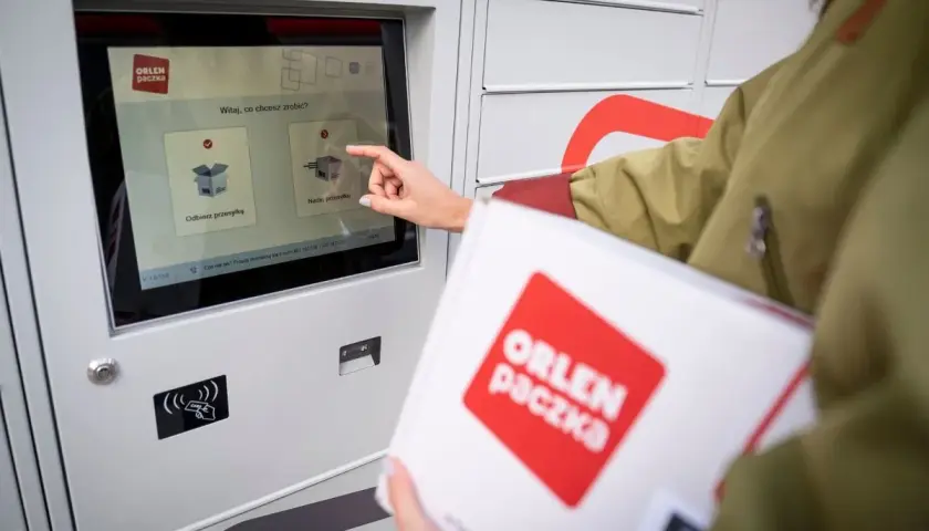Dłoń dotyka ekranu automatu paczkowego, wybierając opcję "Nadaj przesyłkę". W ręku trzymana jest biała paczka z logo ORLEN paczka.