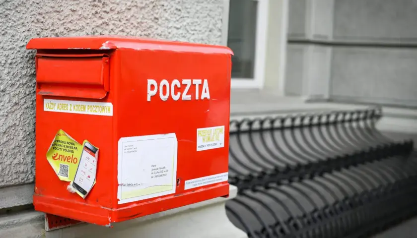 Czerwona skrzynka pocztowa z napisem "POCZTA" i naklejkami. Gotowa na przesyłkę do Wielkiej Brytanii, z adresem i kodem pocztowym.