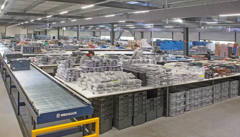 Pracownicy sortują odzież w magazynie. Widać taśmociąg, stoły z zapakowanymi produktami i pojemniki. To pokazuje różne funkcje magazynu.
