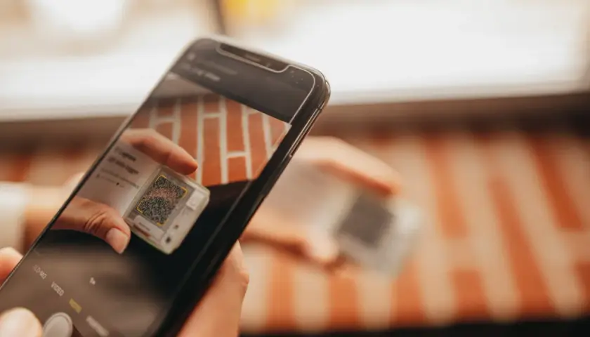 Ręce trzymają smartfon, który skanuje kod QR na małym urządzeniu. To może być część procesu śledzenia przesyłki przez kuriera Hellmann.