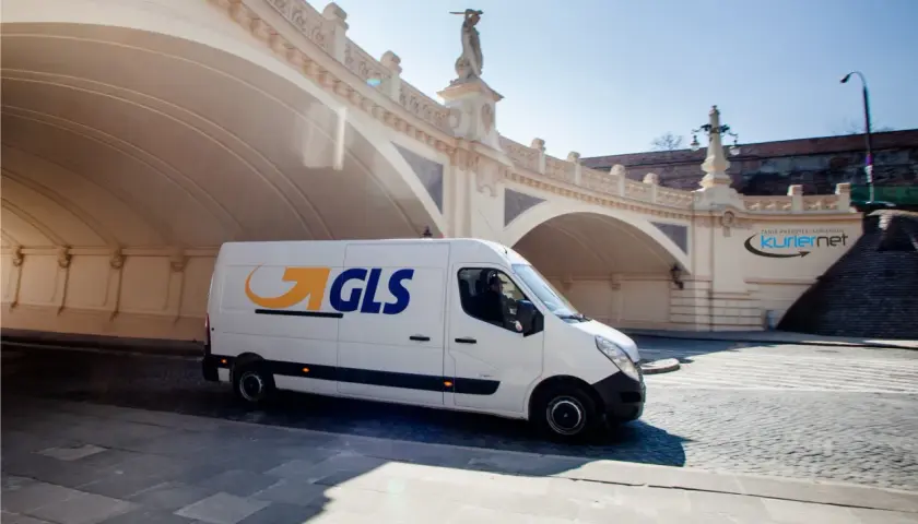 Biały van GLS z logo firmy jedzie pod łukowym mostem, dostarczając paczkę.