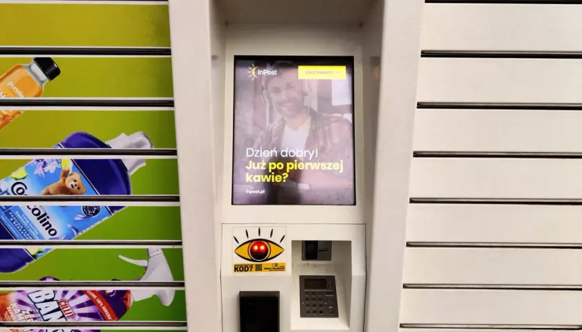 Ekran automatu InPost z uśmiechniętym mężczyzną i pytaniem "Dzień dobry! Już po pierwszej kawie?". Obok widać produkty z półek.