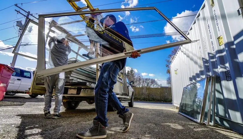 Dwóch mężczyzn zajmuje się transportem okien. Jedno okno jest przenoszone, a inne leżą obok kontenera.