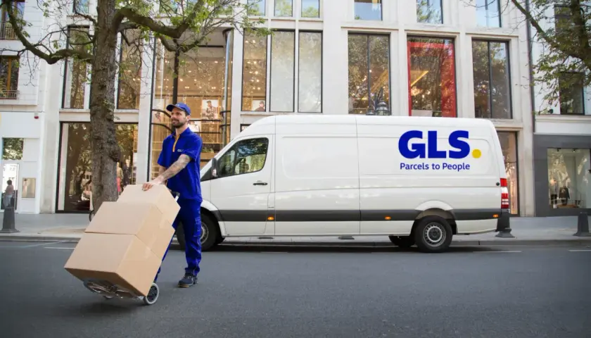 Kurier GLS z paczkami na wózku przed białym vanem. Dowiedz się, jak nadać paczkę z GLS.