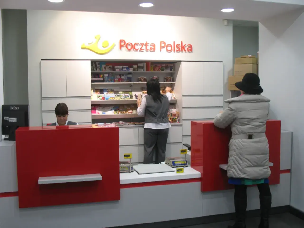 W placówce Poczty Polskiej, pracownica obsługuje klienta. W tle półki z produktami, a obok stos paczek. To miejsce, gdzie realizowane są usługi global expres.