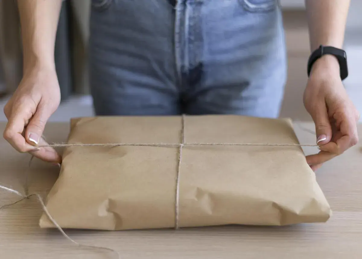 Ręce zawiązują sznurkiem paczkę owiniętą w brązowy papier. Prosty sposób, jak pakować paczki, by dotarły bezpiecznie.