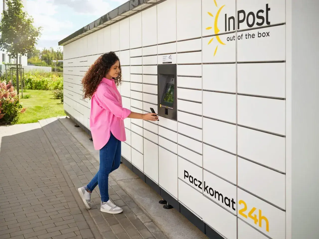 Kobieta z telefonem przy Paczkomacie InPost. Czy z paczkomatu można wysłać paczkę do domu? Tak, to wygodne rozwiązanie.