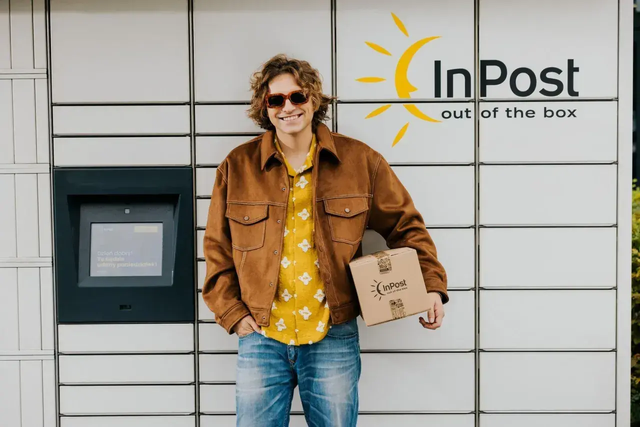 Uśmiechnięty mężczyzna z paczką InPost przy automacie. Sprawdza godziny graniczne InPost, by nadać przesyłkę.
