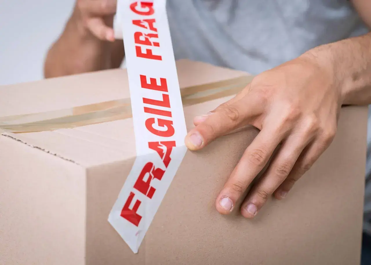Ręce zaklejają karton taśmą z napisem "FRAGILE". Uwaga szkło kurier musi być ostrożny.
