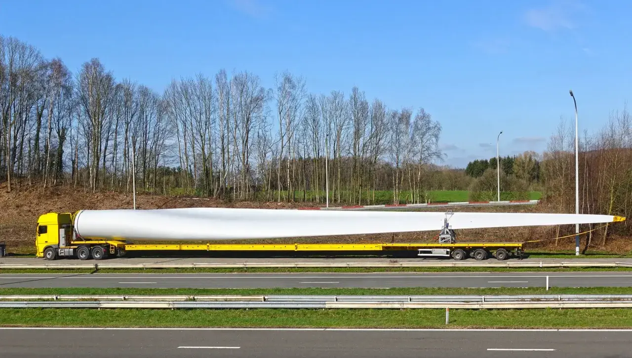 Żółta ciężarówka transportuje ogromne łopaty wiatraka, które są **ładunkami nienormatywnymi**. Długi, biały element spoczywa na specjalnej platformie.