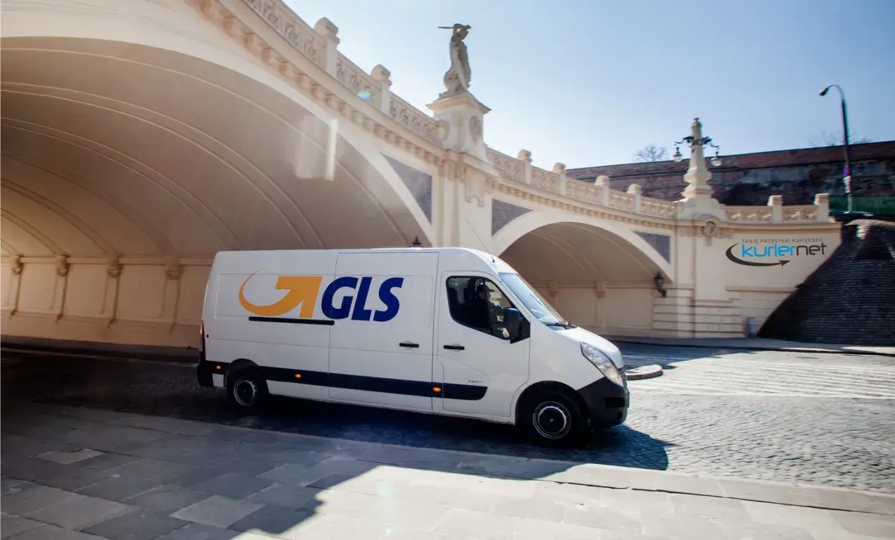 Biały van GLS z logo firmy jedzie pod łukowym mostem, dostarczając paczkę.