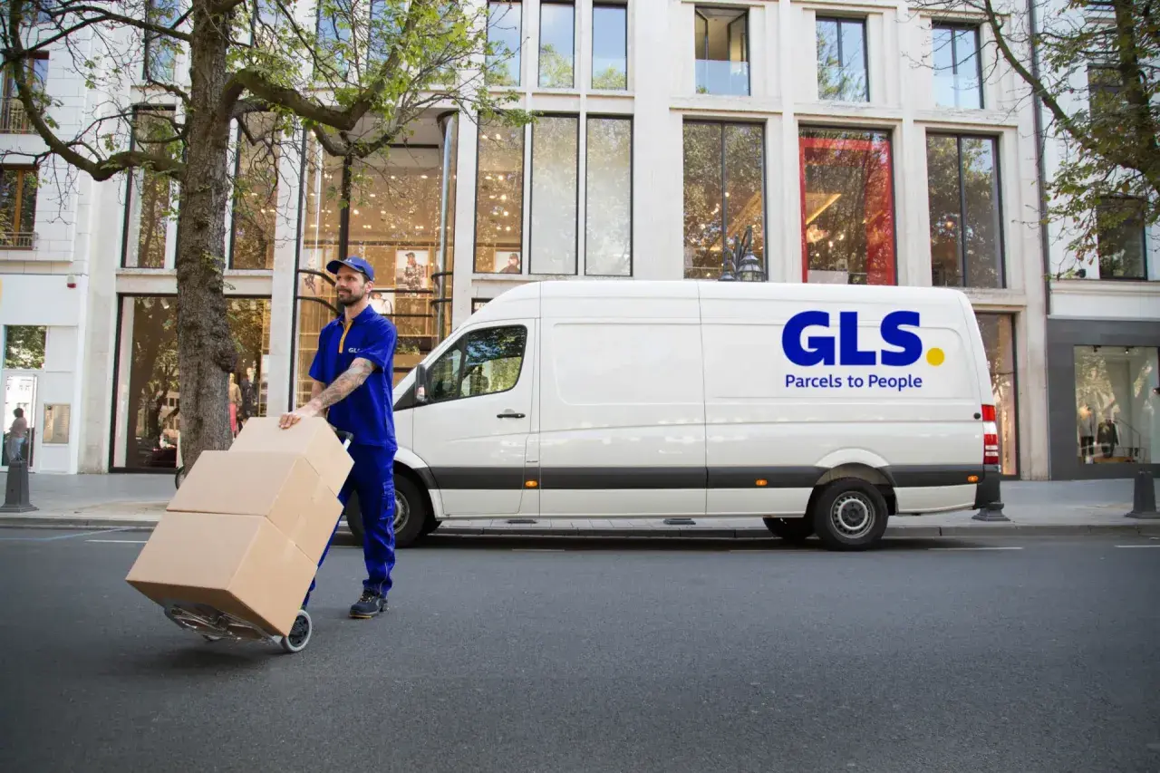 Kurier GLS z paczkami na wózku przed białym vanem. Dowiedz się, jak nadać paczkę z GLS.