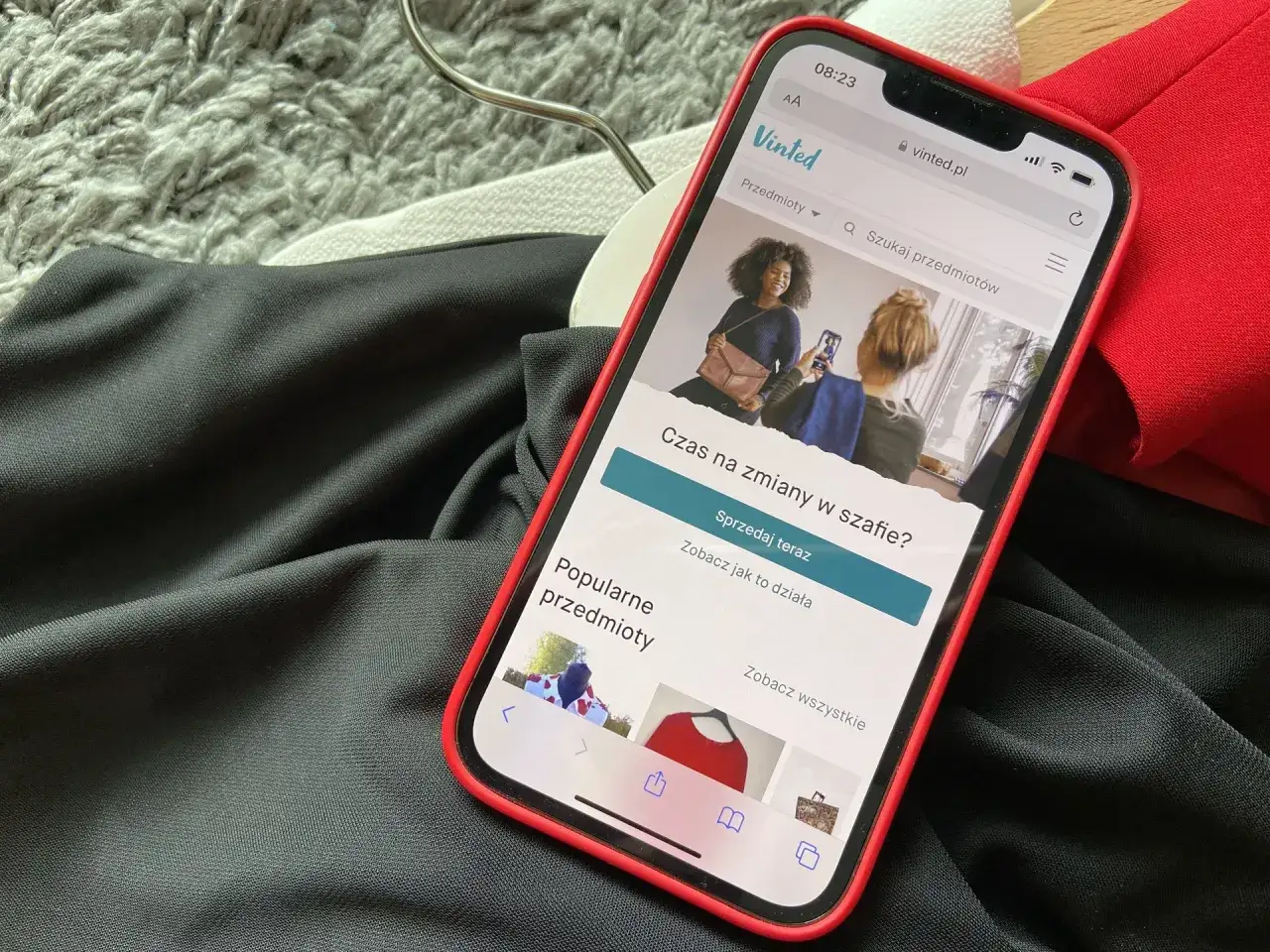 Telefon z aplikacją Vinted na ekranie, obok ubrania. Chcesz wiedzieć, jak zrobić zwrot na Vinted? Sprawdź aplikację!